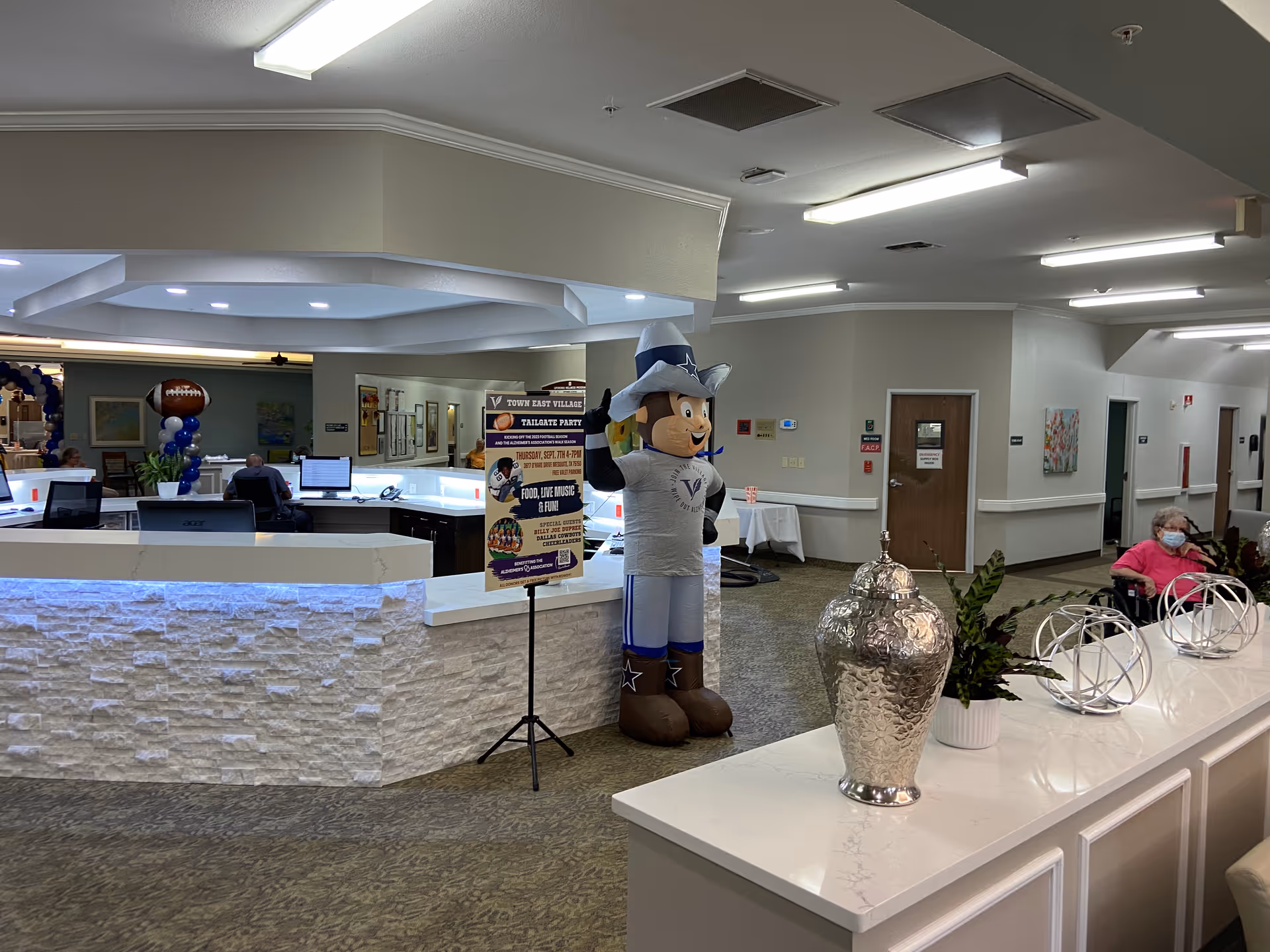 Lobby reception area with a front desk, an inflatable mascot, decorative objects on a countertop, and a seated masked resident.