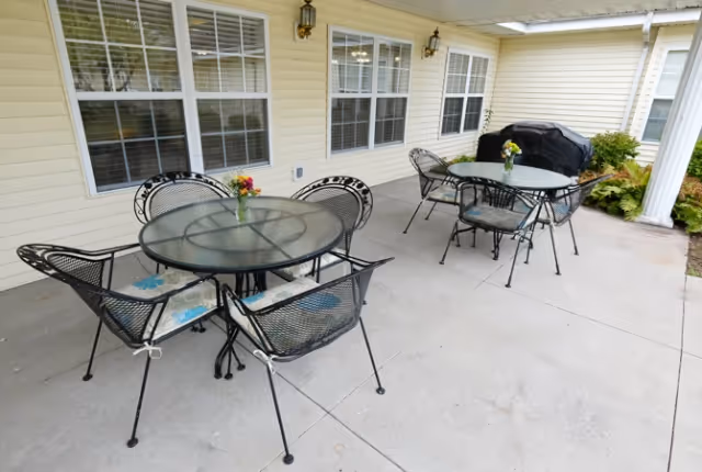 Covered outdoor patio with two round glass-top tables, metal chairs with cushions, small vases of flowers, a grill, and windows on a cream-colored building.