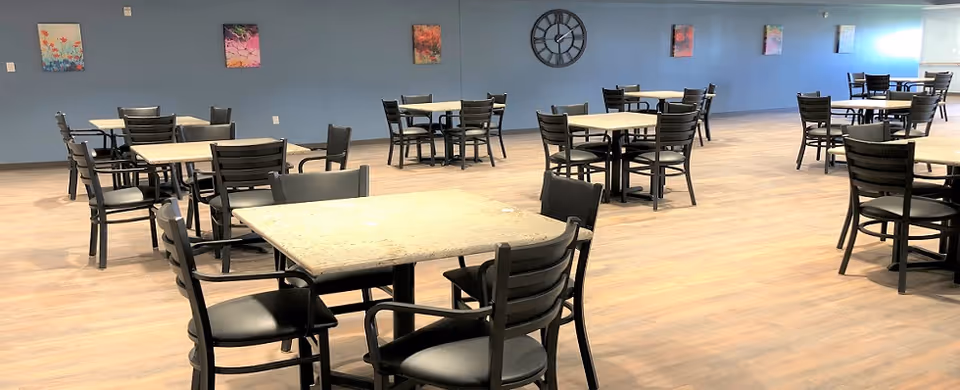 A spacious dining room with multiple square tables and black chairs arranged neatly on a wooden floor. The walls are painted blue and decorated with several colorful paintings and a large black clock.