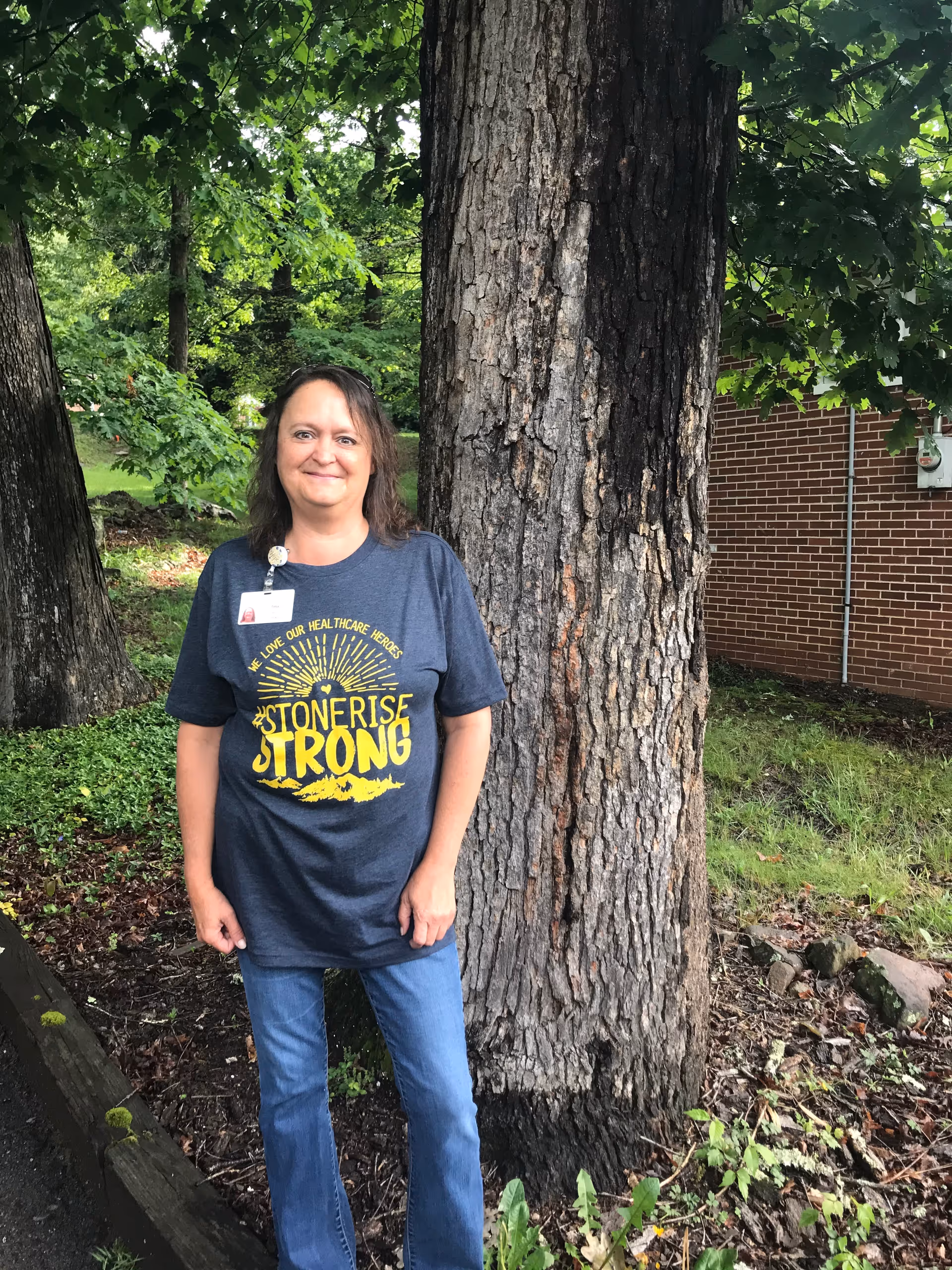 A woman standing outdoors next to a large tree trunk with green foliage and a brick building in the background. She is wearing a dark blue t-shirt with yellow text that reads 'STONE RISE STRONG' and blue jeans. She has a badge clipped to her shirt.
