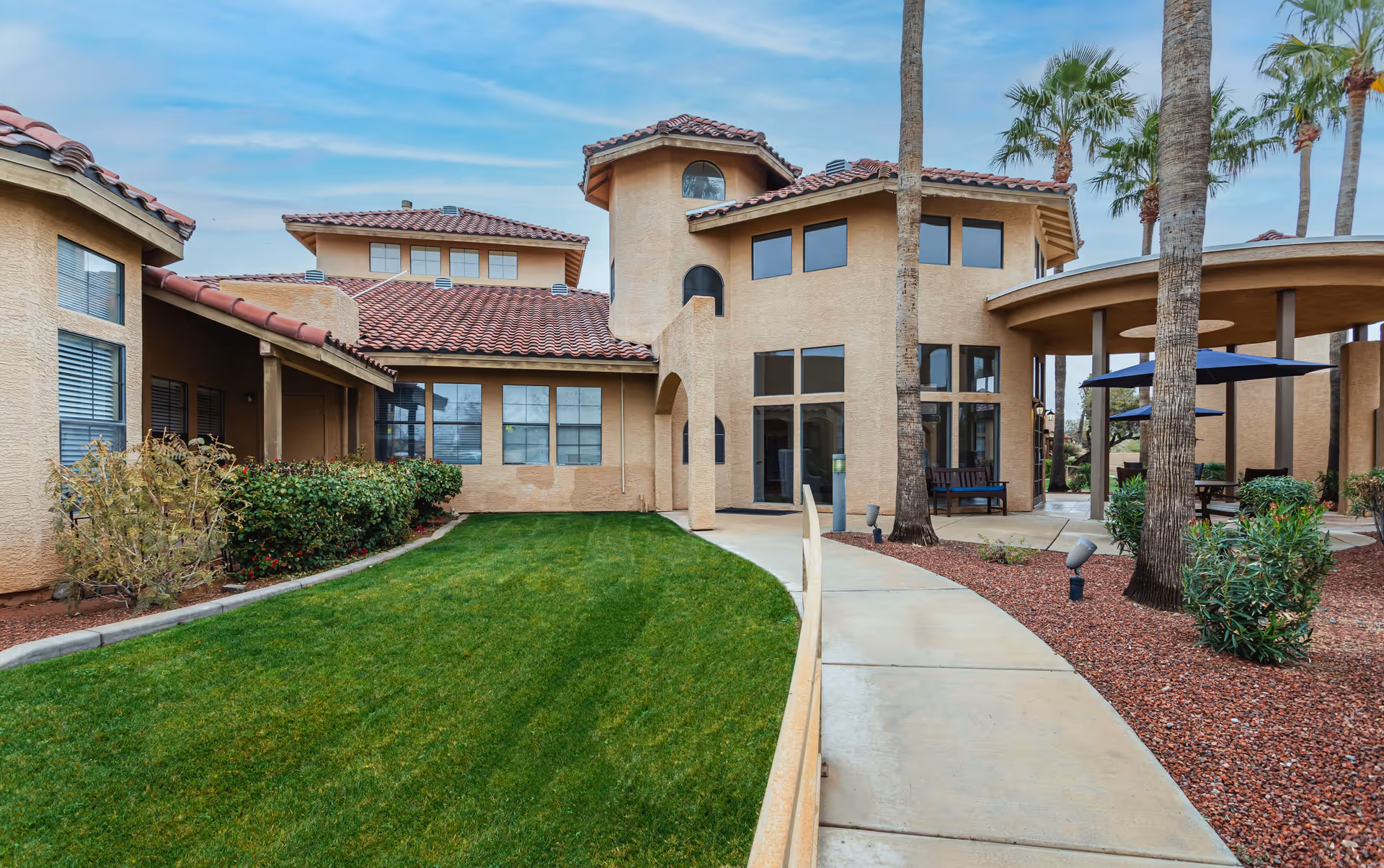 Exterior view of a senior living facility with beige stucco walls and red tile roofs. The building features large windows and a covered patio area with blue umbrellas and benches. There is a curved concrete walkway bordered by green grass on one side and red mulch with palm trees and shrubs on the other side. The sky is partly cloudy.