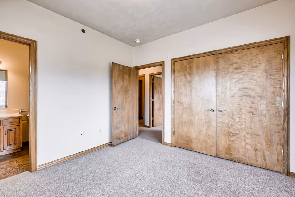 Empty bedroom with beige carpet, light-colored walls, wooden trim, and wooden doors including a double-door closet. A bathroom with wooden cabinetry and a mirror is visible through an open door on the left.