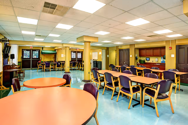 Spacious communal dining/activity room with round and rectangular tables and chairs, yellow support columns, and a kitchenette along the back wall.