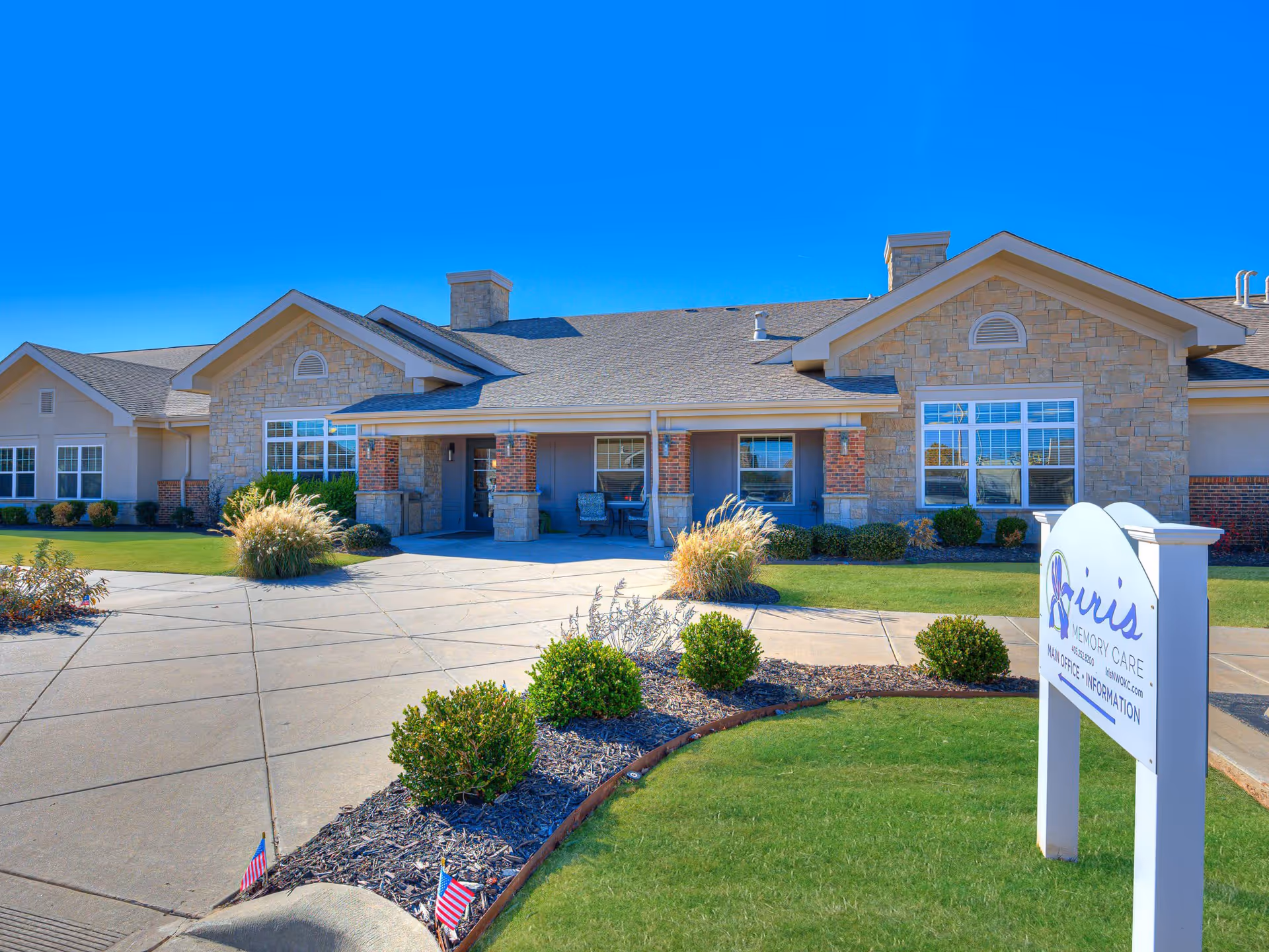 Exterior front view of Iris Memory Care Tulsa facility with a stone and brick facade, a covered entrance with seating, well-maintained landscaping including bushes and ornamental grasses, and a sign displaying the facility name and information.