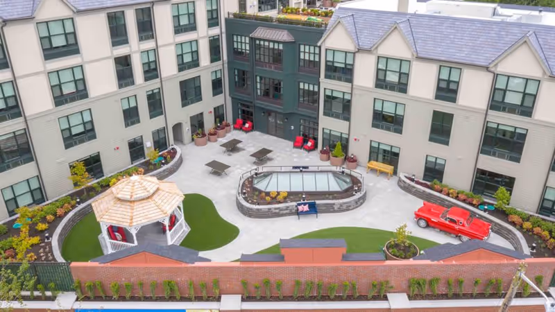 Aerial view of a senior living facility courtyard featuring a gazebo with seating, several tables with chairs, a small garden area, and a red vintage-style car parked near the building. The courtyard is surrounded by a three-story building with many windows and landscaped greenery along the perimeter.