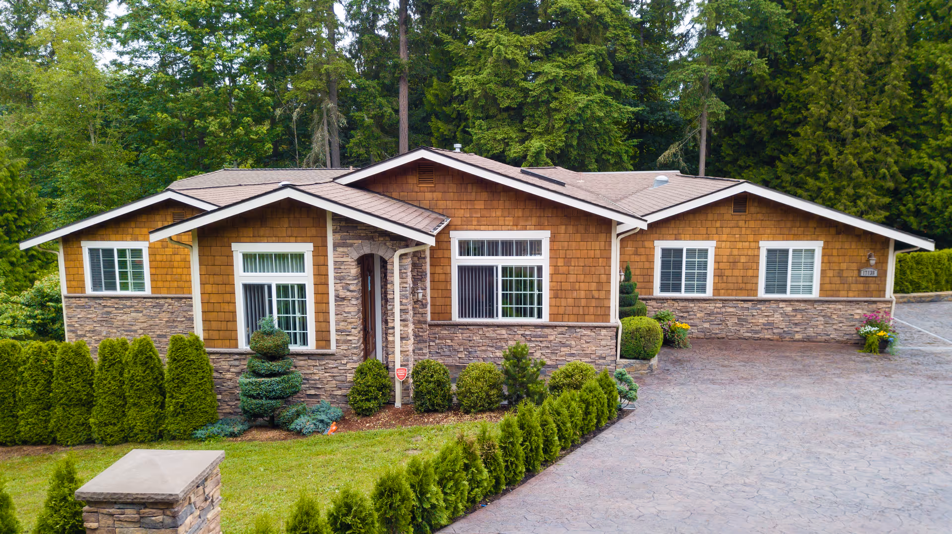 Single-story brown shingle and stone building with a paved driveway and landscaped shrubs in front, set against tall trees.