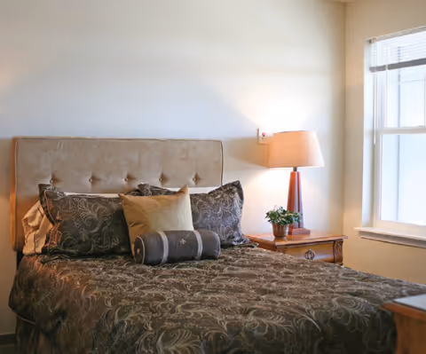 A neatly made bedroom with a tufted headboard, patterned bedspread, decorative pillows, a bedside table with a lamp and a window.