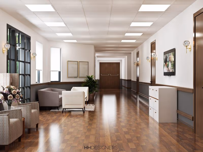 A bright and spacious hallway in a senior living facility with wooden flooring, white walls, and several wooden doors along the right side. The left side features large windows, a seating area with armchairs and a sofa, a potted plant, and framed artwork on the wall. Wall-mounted lights and a flat-screen TV are also visible.