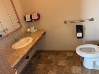 A clean and simple bathroom with a white toilet, a countertop with a sink, a mirror above the sink, a towel rack with two folded towels, a tissue box, and a grab bar next to the toilet. The floor has square tiles in various shades of brown.