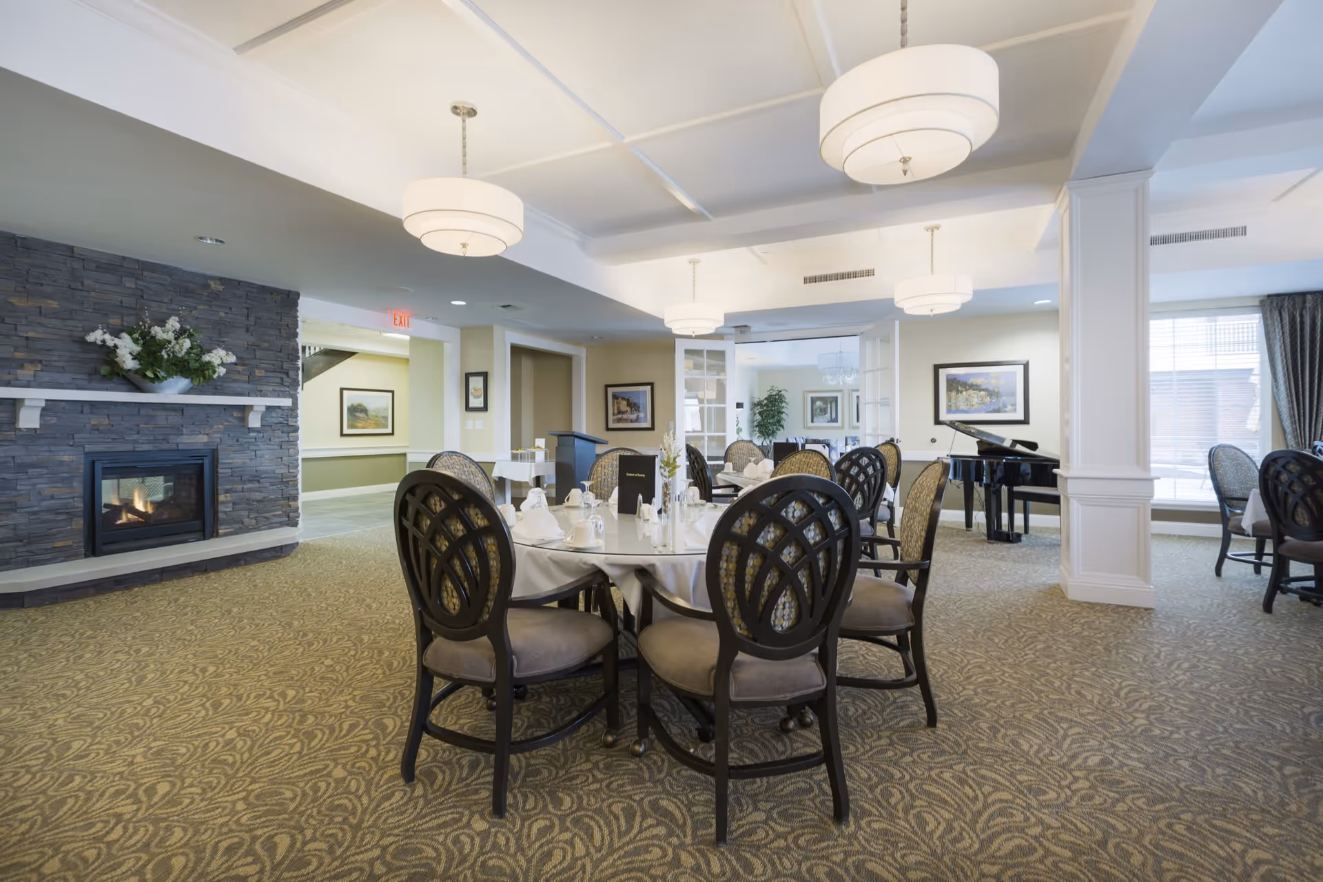 A spacious dining room with round tables set for a meal, featuring elegant chairs with cushioned seats. The room has a patterned carpet, a stone fireplace with a floral arrangement on the mantel, and several hanging light fixtures. In the background, there is a black grand piano near large windows with curtains, and framed artwork on the walls.