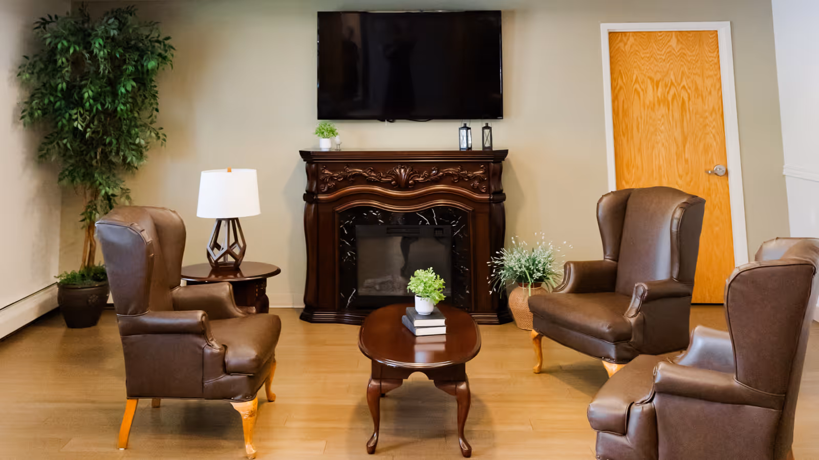 A cozy living room area with four brown leather armchairs arranged around a wooden coffee table with a small plant and books on it. There is a wooden side table with a lamp, a large decorative fireplace with intricate carvings, a flat-screen TV mounted above it, and two potted plants in the corners. The room has light-colored walls and wooden flooring.