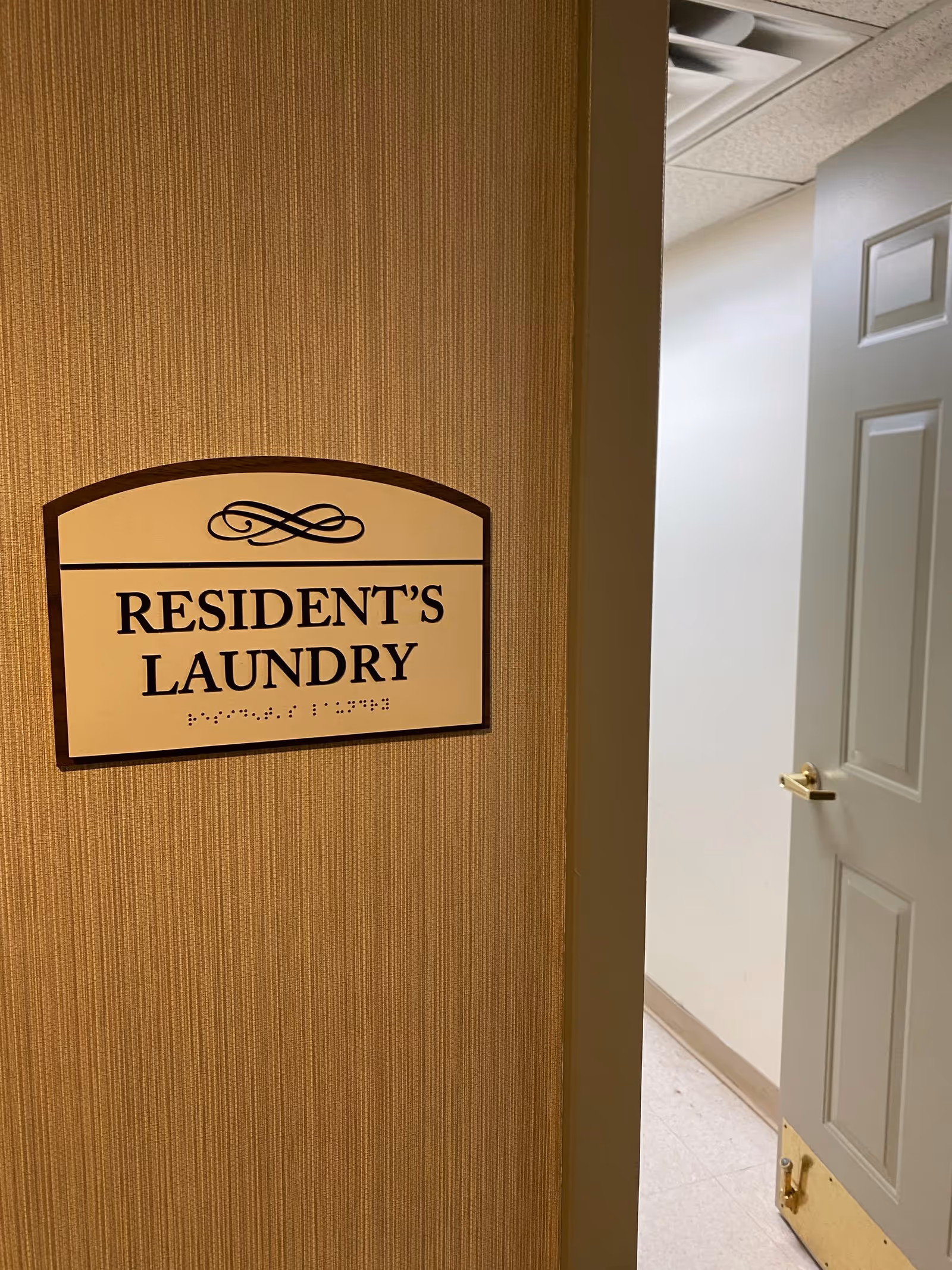 A beige textured wall with a sign that reads 'RESIDENT'S LAUNDRY' in black letters with braille underneath. To the right, a partially open gray door reveals a white hallway with a tiled floor and a ceiling vent.