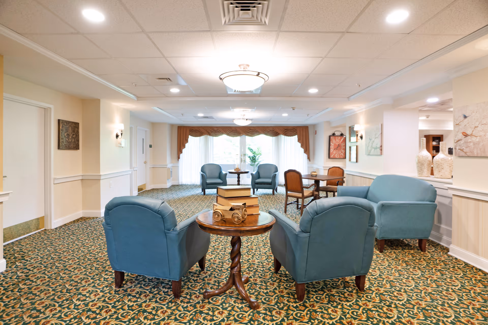 A bright seating lounge with teal armchairs arranged around small wooden tables on a patterned carpet in a senior living facility.