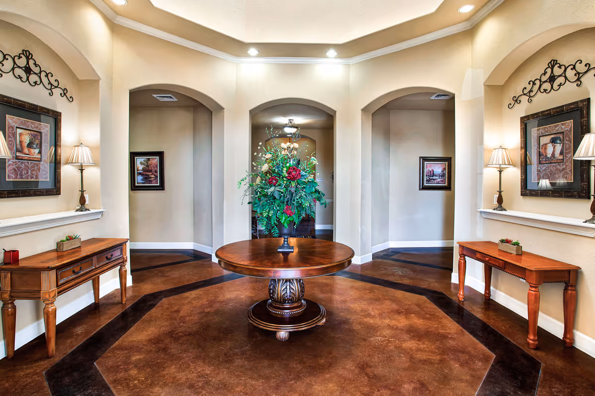 An elegant interior hallway with a round wooden table in the center holding a large floral arrangement. The walls are beige with three arched doorways in the background. On either side of the hallway are wooden console tables with lamps, framed artwork, and small decorative items. The floor has a polished brown finish with a darker border design.