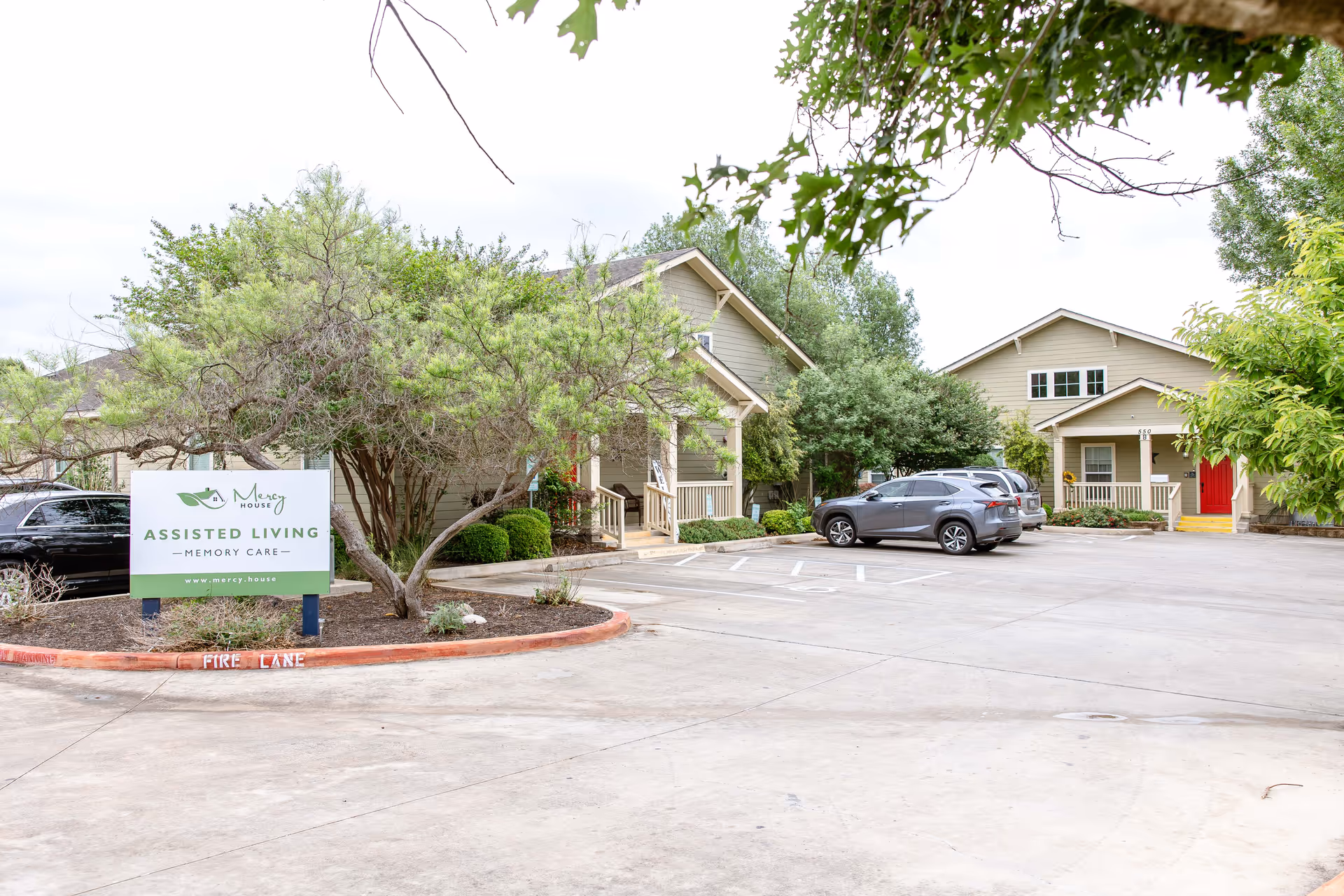 Exterior view of Mercy House New Braunfels assisted living and memory care facility showing a parking lot with several cars, trees, and two beige buildings with porches. A sign in the foreground reads 'Mercy House Assisted Living Memory Care www.mercy.house'.
