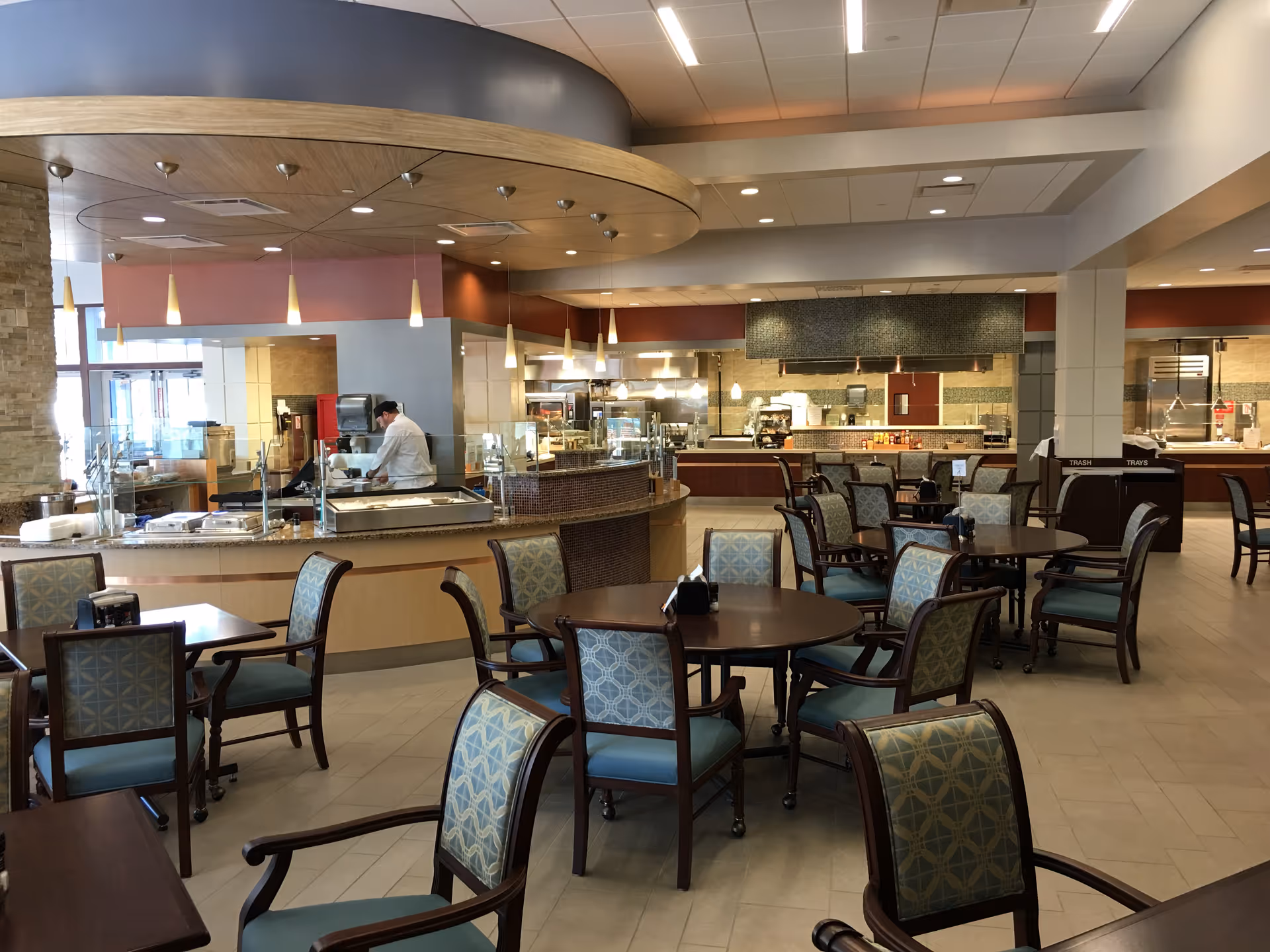 A spacious dining area with multiple round tables and cushioned chairs arranged neatly. In the background, there is a food service counter with a chef preparing food behind a glass partition. The ceiling has modern pendant lights and a curved wooden accent. The overall setting is clean and well-lit.