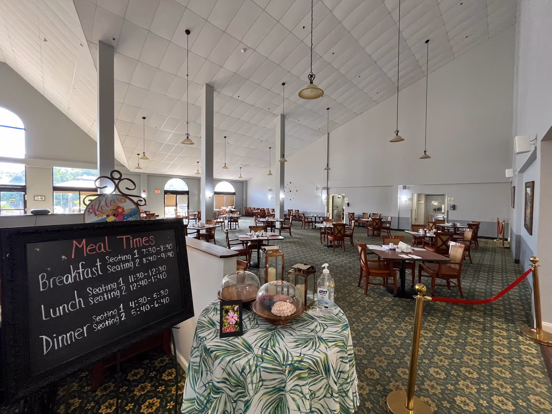 A spacious dining room with multiple wooden tables and chairs arranged neatly. The room has a high, white paneled ceiling with hanging light fixtures. Near the entrance, there is a small table covered with a green and white leaf-patterned cloth, displaying covered plates of food, a bottle of hand sanitizer, and a small framed picture. A blackboard sign lists meal times for breakfast, lunch, and dinner. Large windows allow natural light to fill the room.