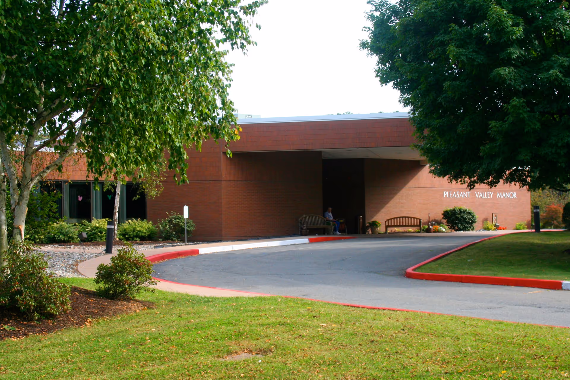 Exterior view of Pleasant Valley Manor Inc, showing a single-story brick building with a covered entrance. There are benches near the entrance, a curved driveway with red and white painted curbs, and green trees and grass surrounding the area.