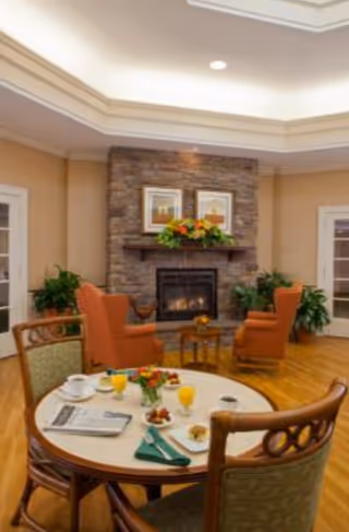 A cozy living room area in Glenmere Assisted Living featuring a stone fireplace with a mantel decorated with flowers and framed pictures above it. Two orange armchairs face the fireplace, surrounded by green plants. In the foreground, a round table is set with breakfast items including orange juice, coffee cups, plates with food, and a newspaper. The room has warm lighting and wooden flooring.
