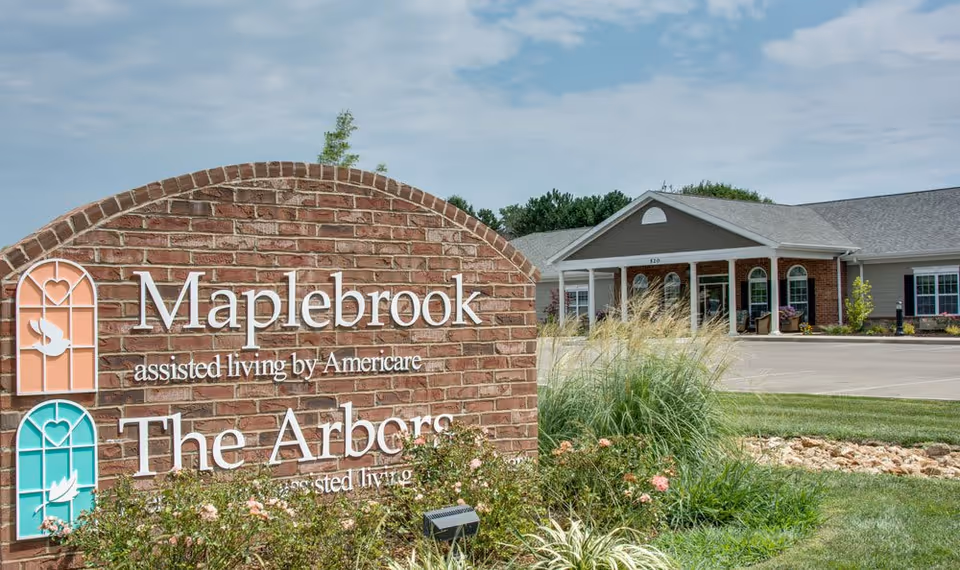 Brick entrance sign reading 'Maplebrook assisted living by Americare / The Arbors' with landscaped grounds and the facility's single-story entrance building in the background.