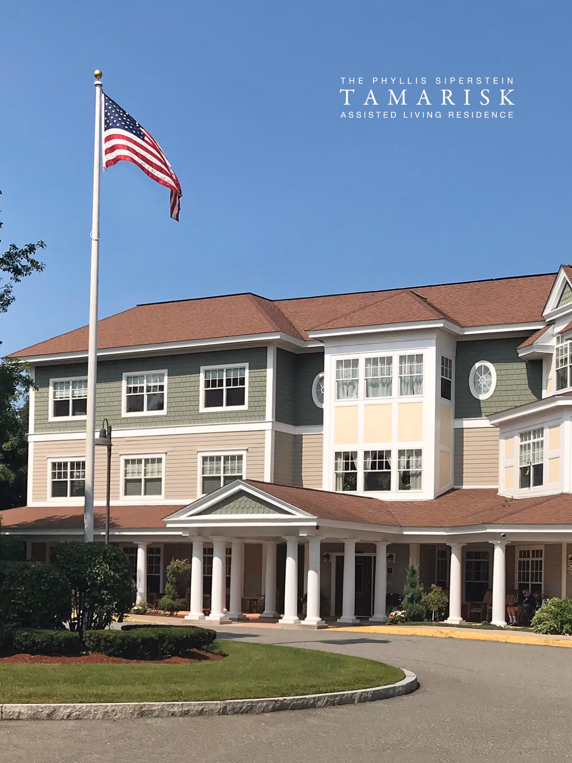 Exterior view of Tamarisk Senior Living residence showing a multi-story building with beige and green siding, white columns at the entrance, and an American flag on a flagpole in front under a clear blue sky.