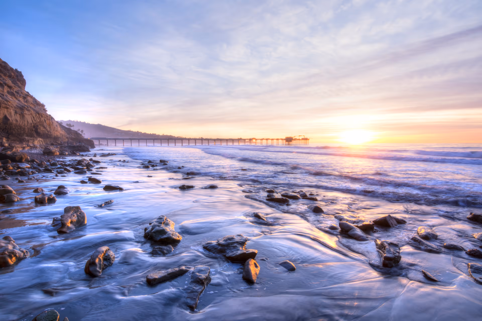A rocky beach at sunset with waves gently washing over the shore. In the background, there is a long pier extending into the ocean under a partly cloudy sky with the sun near the horizon.