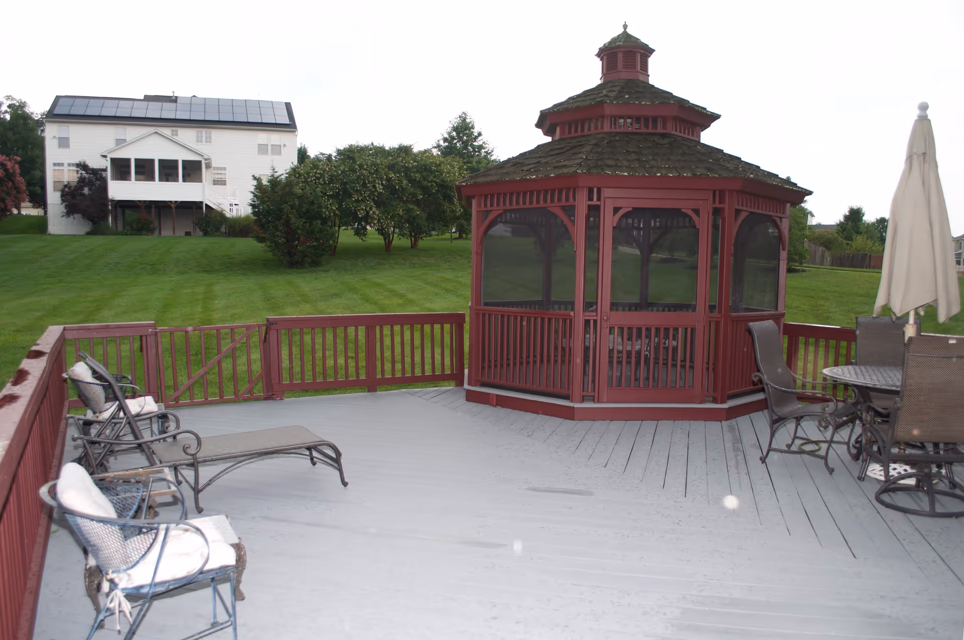 Outdoor patio area with a red wooden gazebo, several metal chairs with cushions, a lounge chair, a round table with chairs, and a closed umbrella. The patio overlooks a large grassy lawn with trees and a white house with solar panels in the background.
