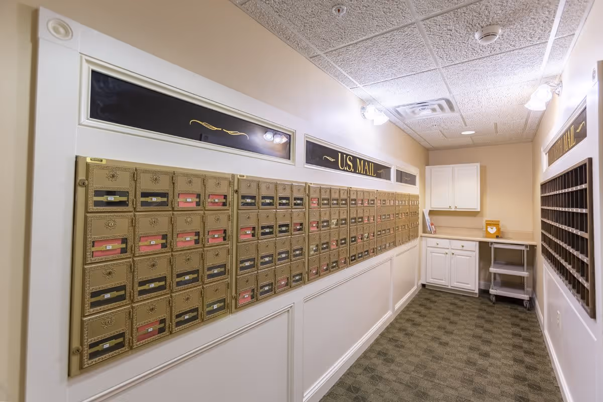 A mailroom with multiple rows of gold-colored mailboxes labeled U.S. Mail on the left wall, a set of white cabinets and a small rolling cart at the end of the room, and a wooden cubby-style mail organizer on the right wall. The room has beige walls, a patterned carpet, and a drop ceiling with fluorescent lights.