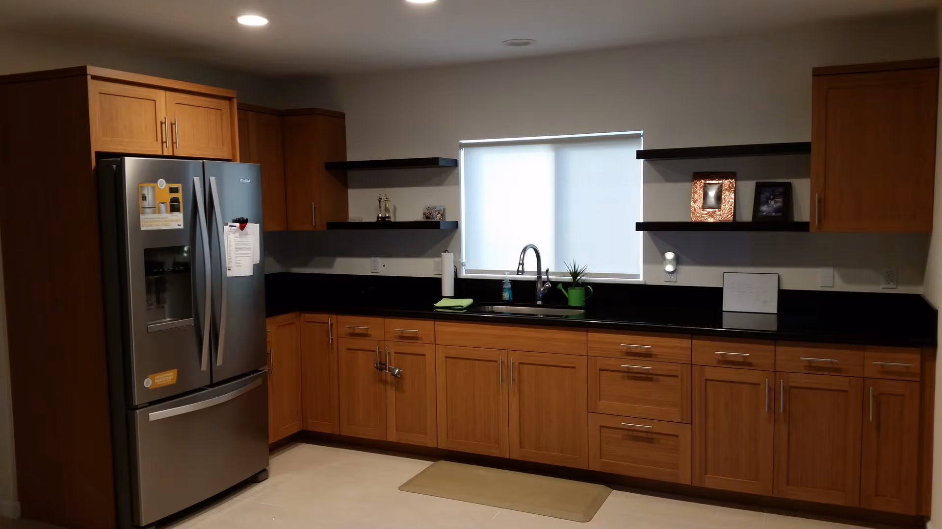 Modern kitchen with wooden cabinets, black countertops, a stainless steel refrigerator, and a sink under a window.