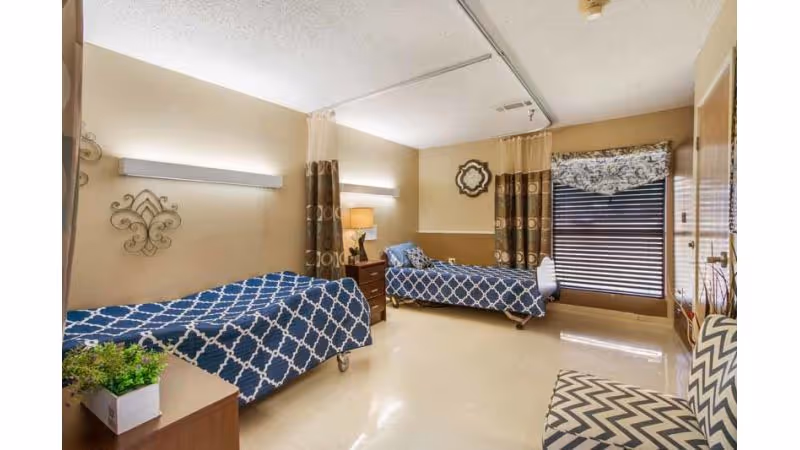 A bright and clean bedroom in a nursing facility with two single beds covered in blue and white patterned bedspreads. The room has beige walls with decorative wall art, a nightstand with a lamp between the beds, a window with patterned curtains and blinds, and a chevron-patterned chair in the corner.