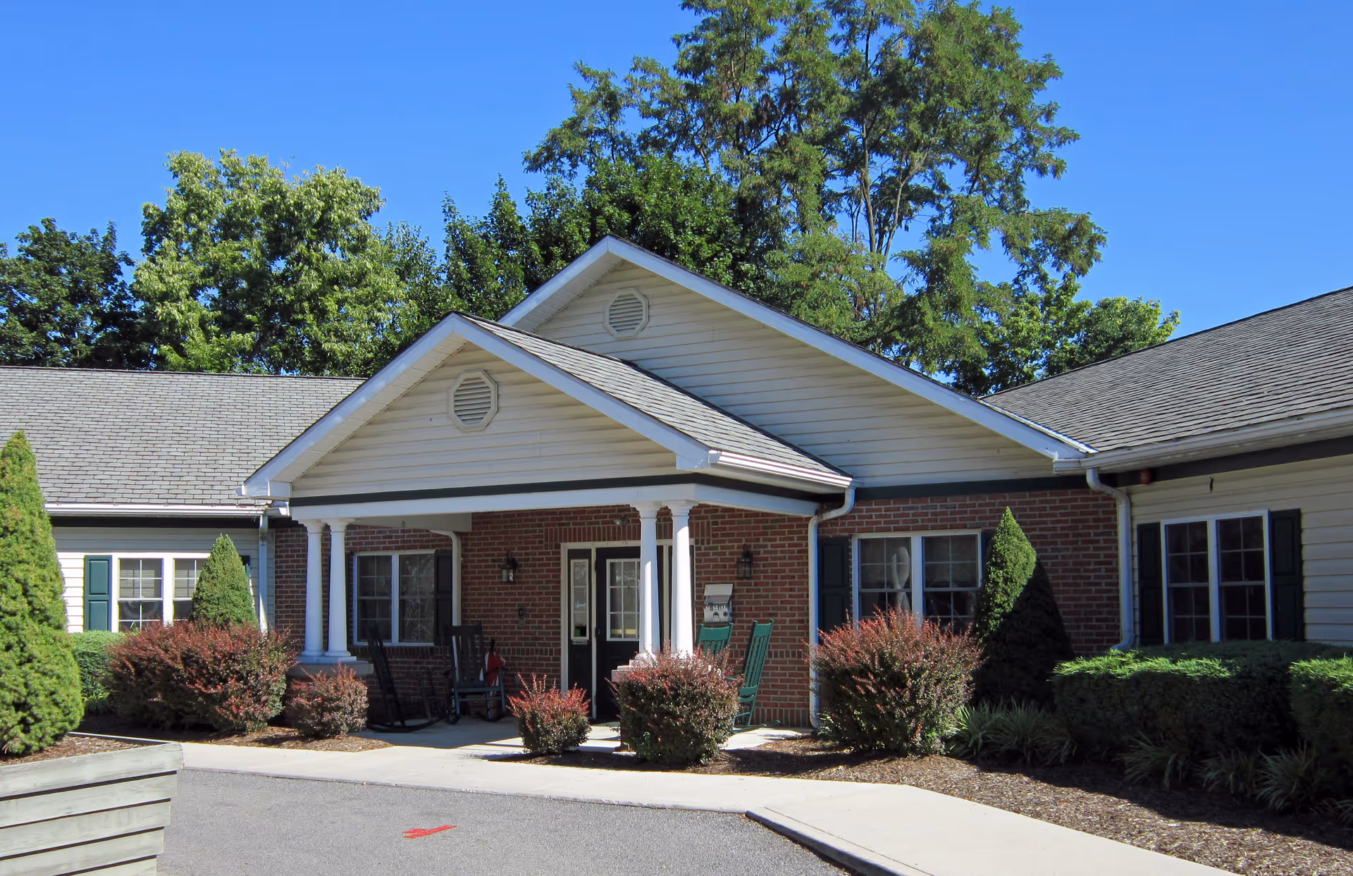 Exterior view of a single-story brick and siding building with a covered porch entrance featuring white columns and rocking chairs. The building is surrounded by neatly trimmed bushes and trees under a clear blue sky.