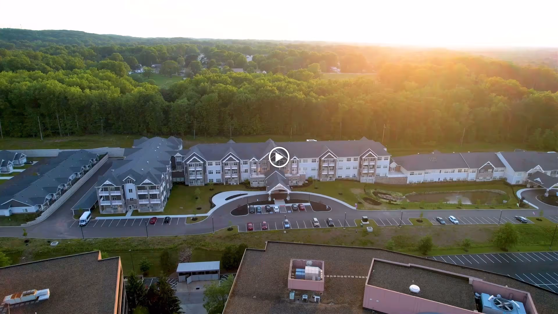 Aerial view of VITALIA Active Adult Community at Rockside, showing a large multi-story residential building with balconies, surrounded by parking lots and green wooded areas in the background during sunset.