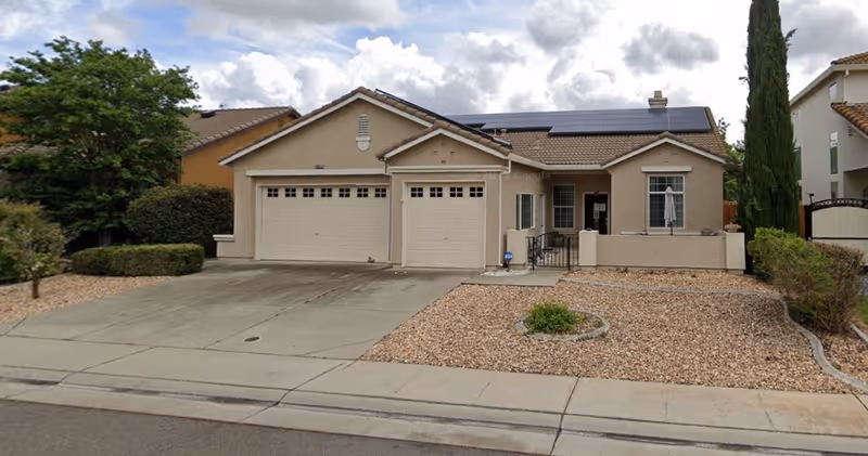 Front exterior view of a single-story house with a two-car garage, beige stucco walls, and a tiled roof with solar panels. The driveway and front yard are covered with gravel and minimal landscaping, including a small circular plant bed and some bushes. The sky is partly cloudy.