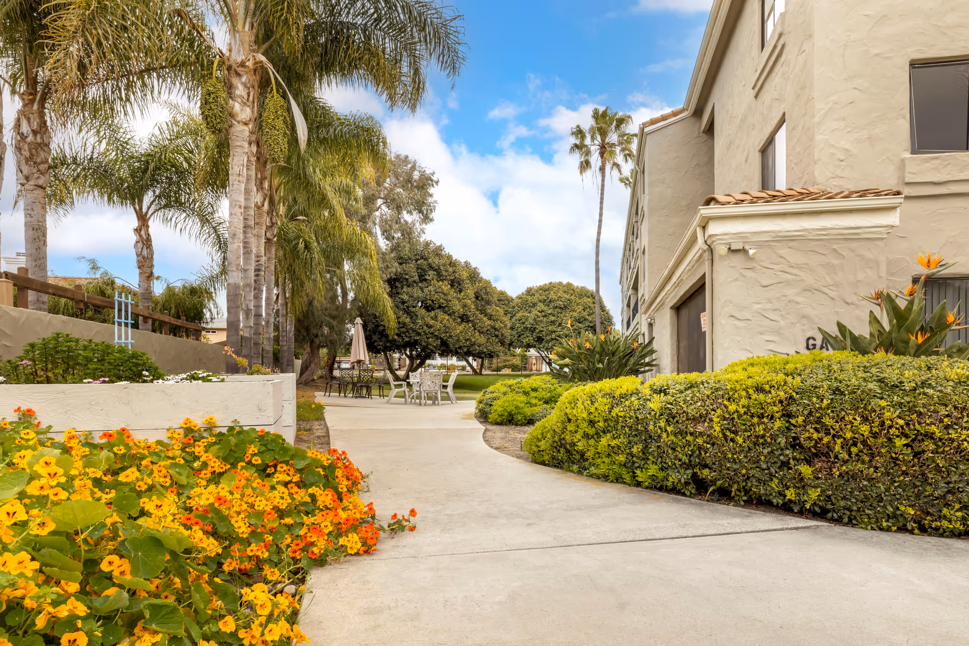 Outdoor pathway at Laguna Estates Senior Living with vibrant yellow and orange flowers on the left, green bushes on the right, palm trees, and a beige building with a tiled roof. There are tables and chairs with umbrellas in the background under a partly cloudy blue sky.