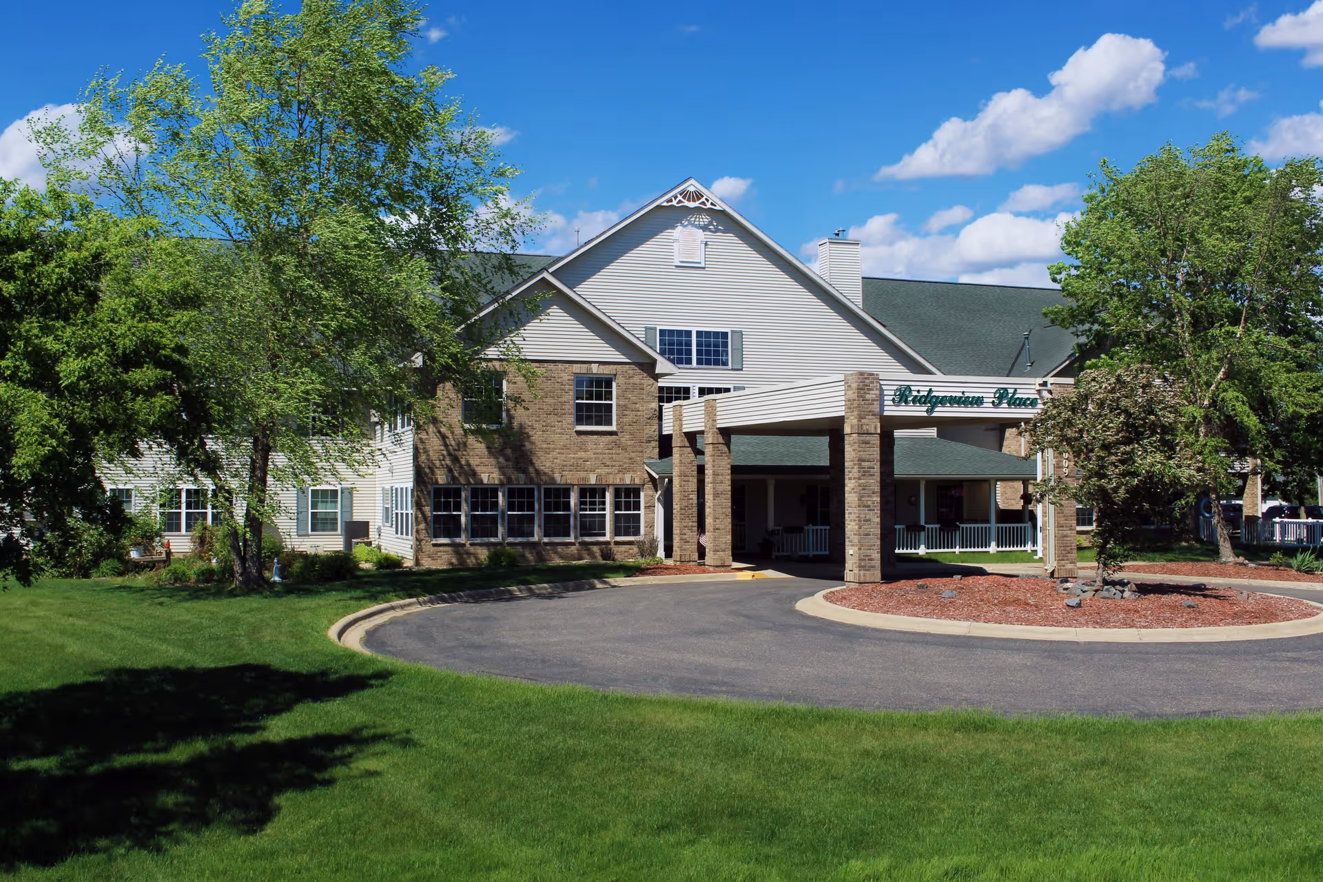 Front exterior of Ridgeview Place Assisted Living with a covered entrance, circular driveway, and trees on a sunny day.