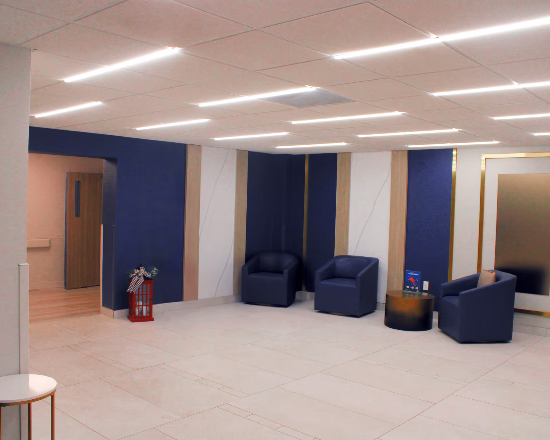 A modern interior seating area with three dark blue armchairs arranged around a small round table. The walls feature vertical panels in blue, white, and beige tones, and the ceiling has linear recessed lighting. A decorative red lantern with a bow is placed near an open doorway leading to another room.