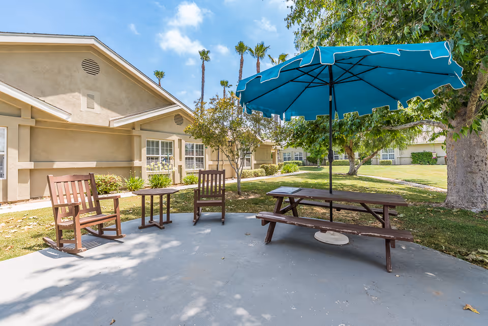 Outdoor courtyard with a picnic table and blue umbrella, wooden chairs, and a single-story beige building in the background.