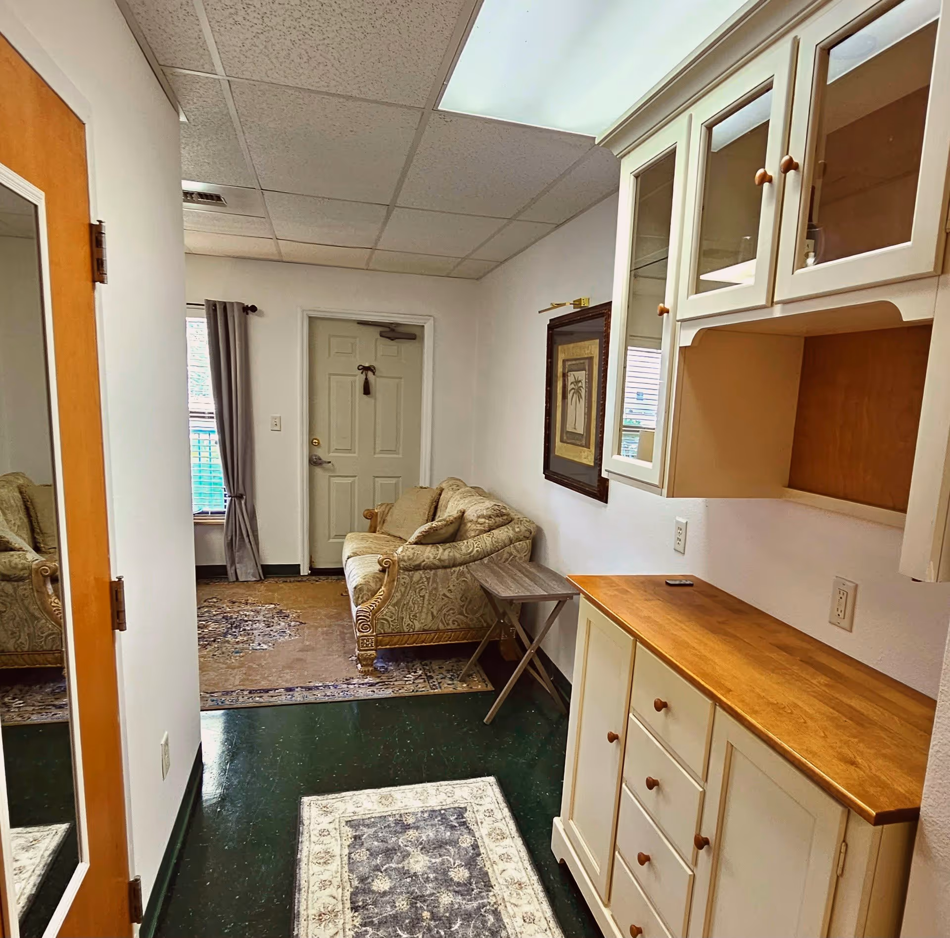 Interior view of a small living area in a senior living facility. The room features a patterned beige sofa with a side table next to it, a framed picture on the wall, and a window with gray curtains. On the right side, there is a white cabinet with glass doors and wooden countertop. The floor has a dark green tile with two area rugs. A door with a mirror on the left side is partially visible.