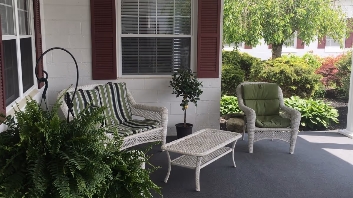 A covered outdoor patio area with white wicker furniture including a loveseat with striped cushions, a chair with a green cushion, and a matching coffee table. There are green plants and bushes surrounding the patio, with a window and red shutters on the building wall behind the furniture.