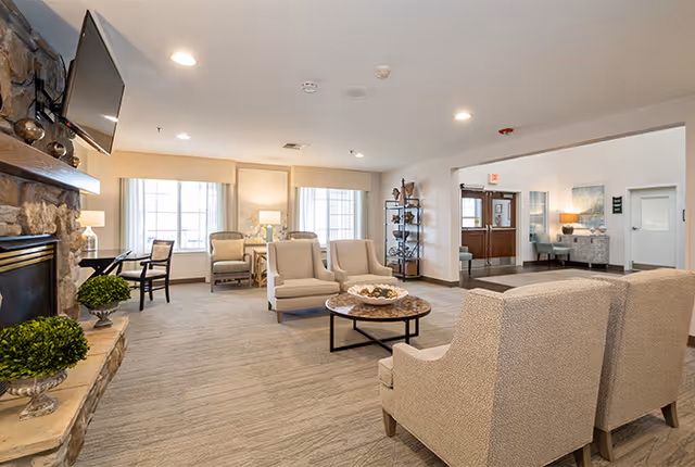 A bright and spacious living room area in a senior living facility with beige upholstered armchairs arranged around a round coffee table. There is a stone fireplace on the left with a mounted flat-screen TV above it. Large windows with light curtains allow natural light to fill the room. The room also features a small desk with a chair near the windows and a shelving unit near the entrance door.