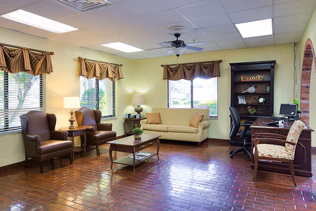 A well-lit living room with three large windows adorned with brown valances. The room features two brown armchairs with a small wooden side table and lamp between them, a beige sofa with two cushions, a wooden coffee table with a plant on it, and a wooden desk with a computer and office chair. There is also a wooden bookshelf with decorative items and the word 'Family' displayed on the top shelf. The floor is covered with reddish-brown tiles, and the ceiling has recessed lighting and a ceiling fan.