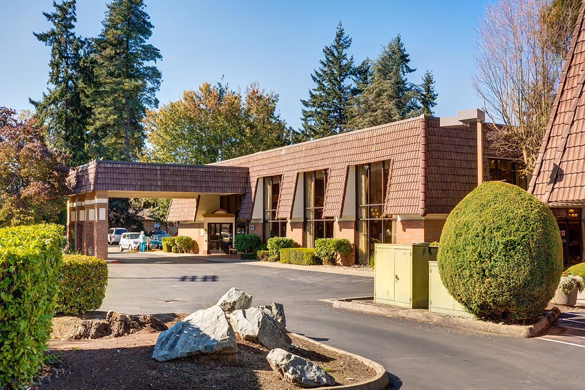 Exterior view of The Rivers at Puyallup facility showing a building with brown shingled roof and large windows, surrounded by trimmed bushes, trees, and rocks under a clear blue sky.