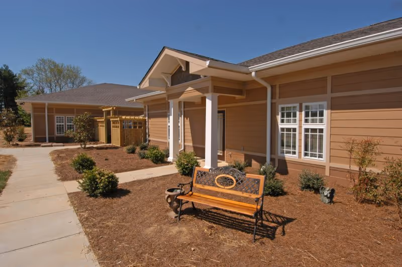 Front exterior of a single-story retirement center with a bench, walkway, and landscaping under a blue sky.
