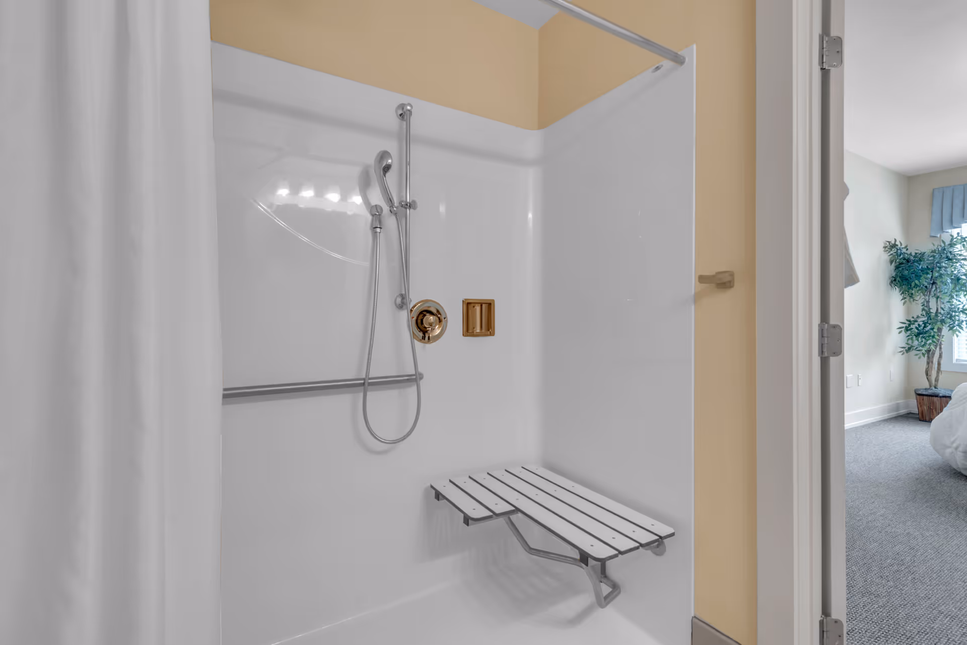 Shower stall with a handheld showerhead, grab bar, and a fold-down bench, view opening into an adjacent bedroom.