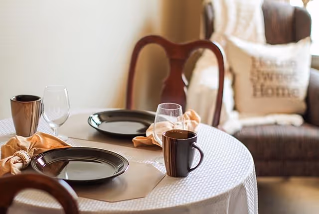 A round dining table set for two with black plates, brown mugs, wine glasses, and beige cloth napkins. In the background, there is a cushioned chair with a pillow that reads 'Home Sweet Home.'