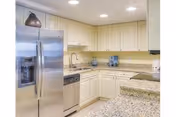 Modern kitchen with cream-colored cabinets, granite countertops, stainless steel refrigerator, dishwasher, and built-in stove. The kitchen is well-lit with recessed ceiling lights and has decorative items on the counter and above the cabinets.