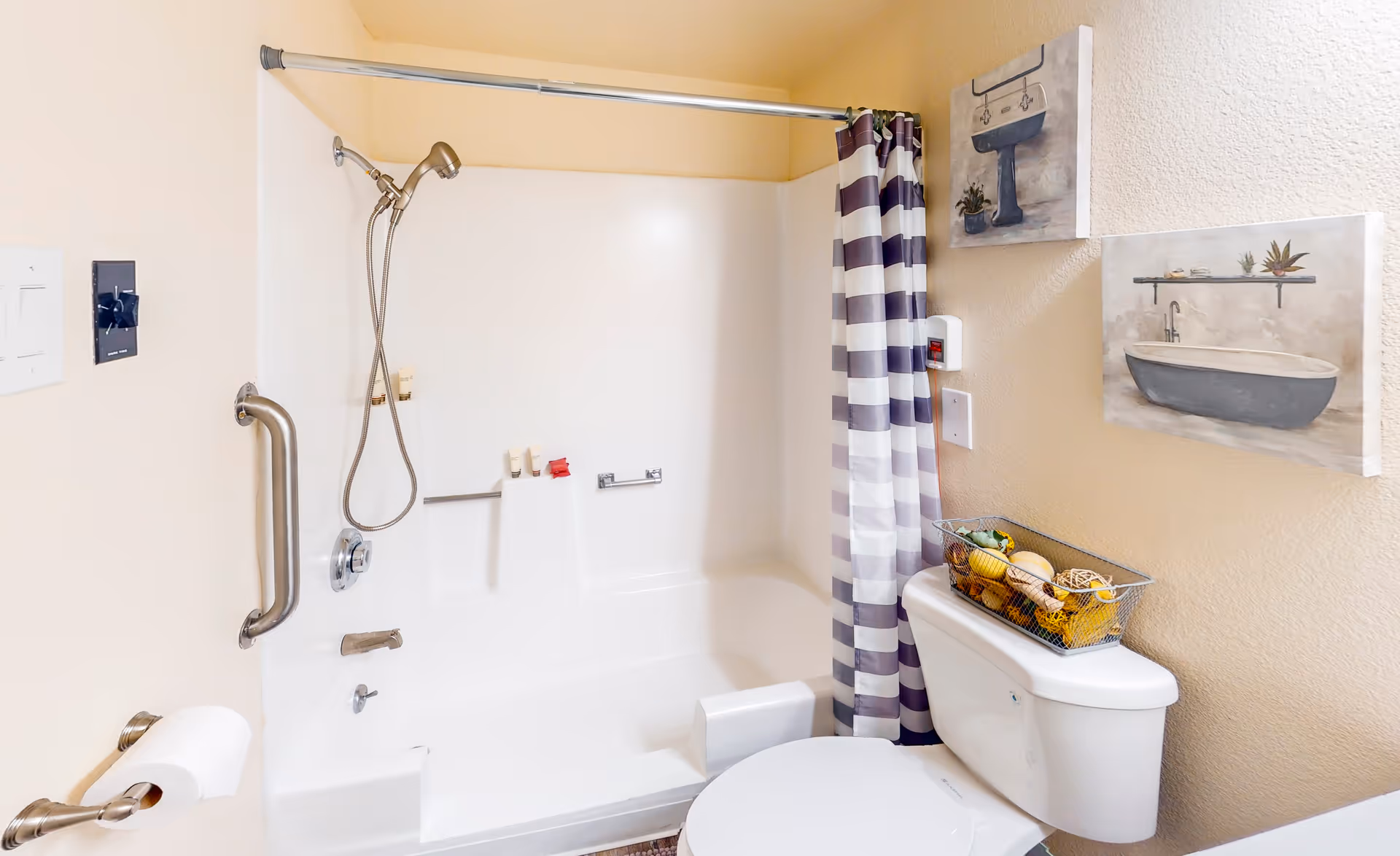 A clean bathroom featuring a white bathtub with a handheld showerhead and a striped shower curtain. There is a grab bar on the wall next to the bathtub, a toilet with a basket of decorative items on the tank, and two framed pictures hanging on the wall above the toilet.