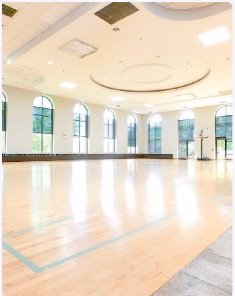A spacious indoor gymnasium with polished wooden floors and multiple large arched windows letting in natural light. The ceiling features recessed lighting and decorative circular molding. A basketball hoop is visible in the far corner.