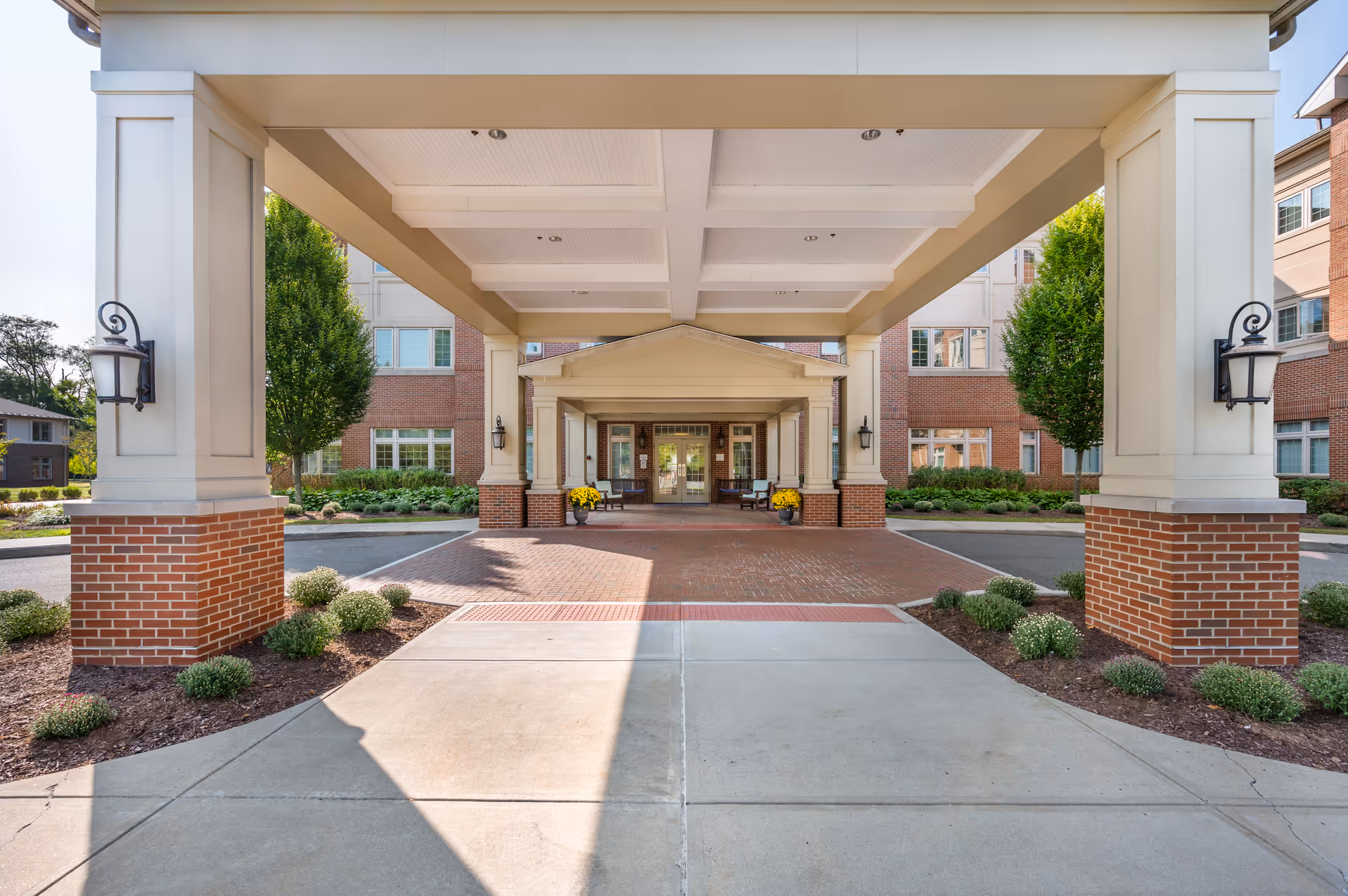 Covered entrance to a senior living facility with brick pillars and beige columns, outdoor lamps on the pillars, potted yellow flowers near the entrance, and a brick building with multiple windows in the background.
