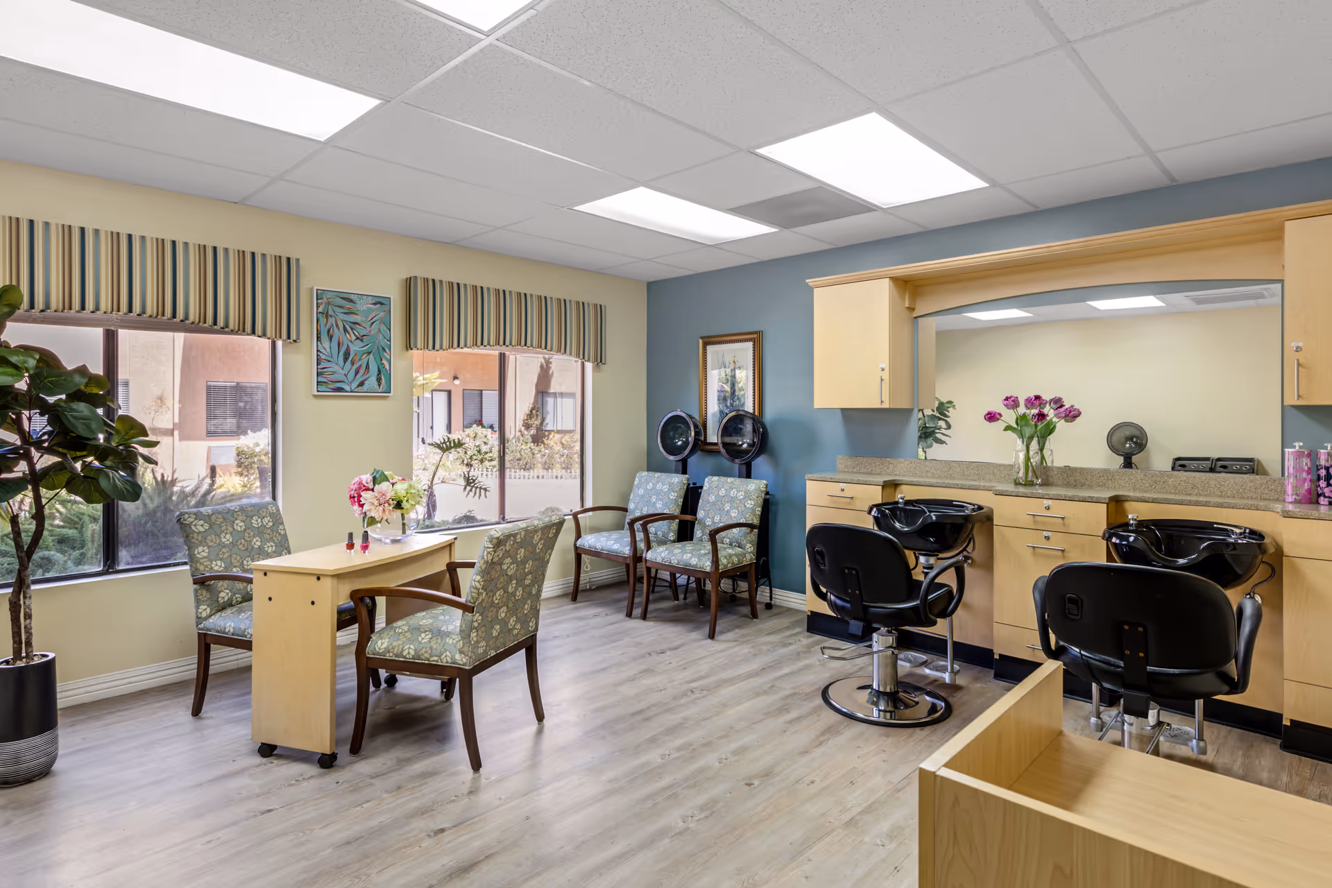 A bright and clean salon area in a senior living facility with two black salon chairs in front of a large mirror and countertop with cabinets. There are two hair drying stations against a blue accent wall. A small table with four patterned chairs is positioned near two large windows with striped valances. The room has light wood flooring and ceiling fluorescent lights.